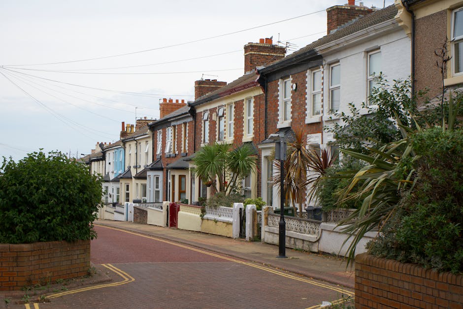 A row of terraced houses along a quiet street, each with small front gardens featuring a variety of plants and trees. The gardens include trimmed bushes, tropical-style palm trees, and leafy shrubs, w