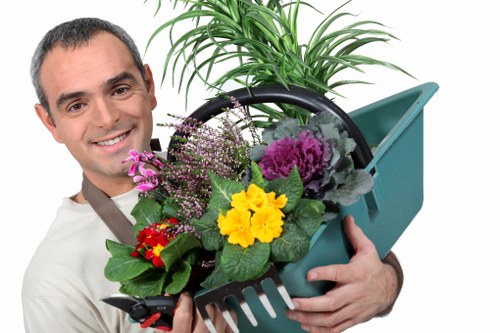 The image depicts a smiling man holding a green plastic garden tool basket filled with a variety of colorful potted plants and flowering flowers, including purple, yellow, and red blossoms. Behind him, there's a lush green garden scene with a well-maintained lawn area, bordered by a hedge or shrubbery, and a background featuring trees and natural outdoor elements. The setting appears to be a landscaped front or back garden in Wimbledon, showcasing healthy, vibrant plant life suitable for professional gardening or landscaping services. The weather is clear and bright, highlighting the natural tones of the plants and outdoor environment, emphasizing the importance of garden maintenance and enhancement services offered by Gardener Wimbledon for local outdoor spaces.